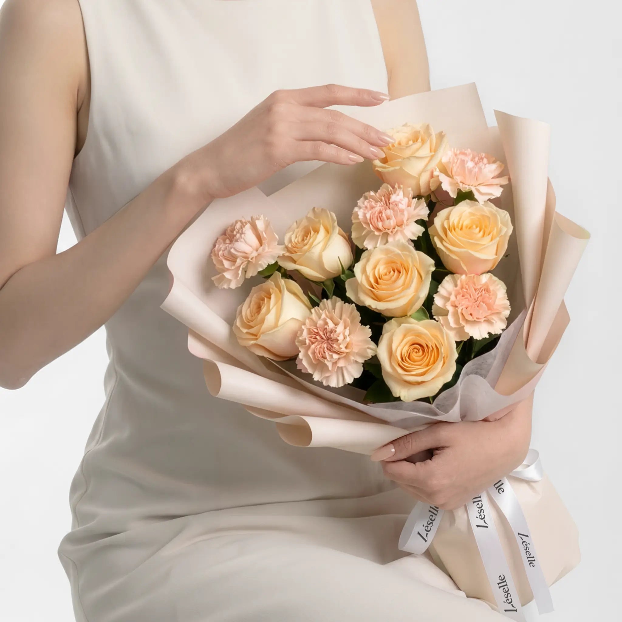 Bouquet of champagne roses and peach pink carnations held by a person in a white dress on a white background